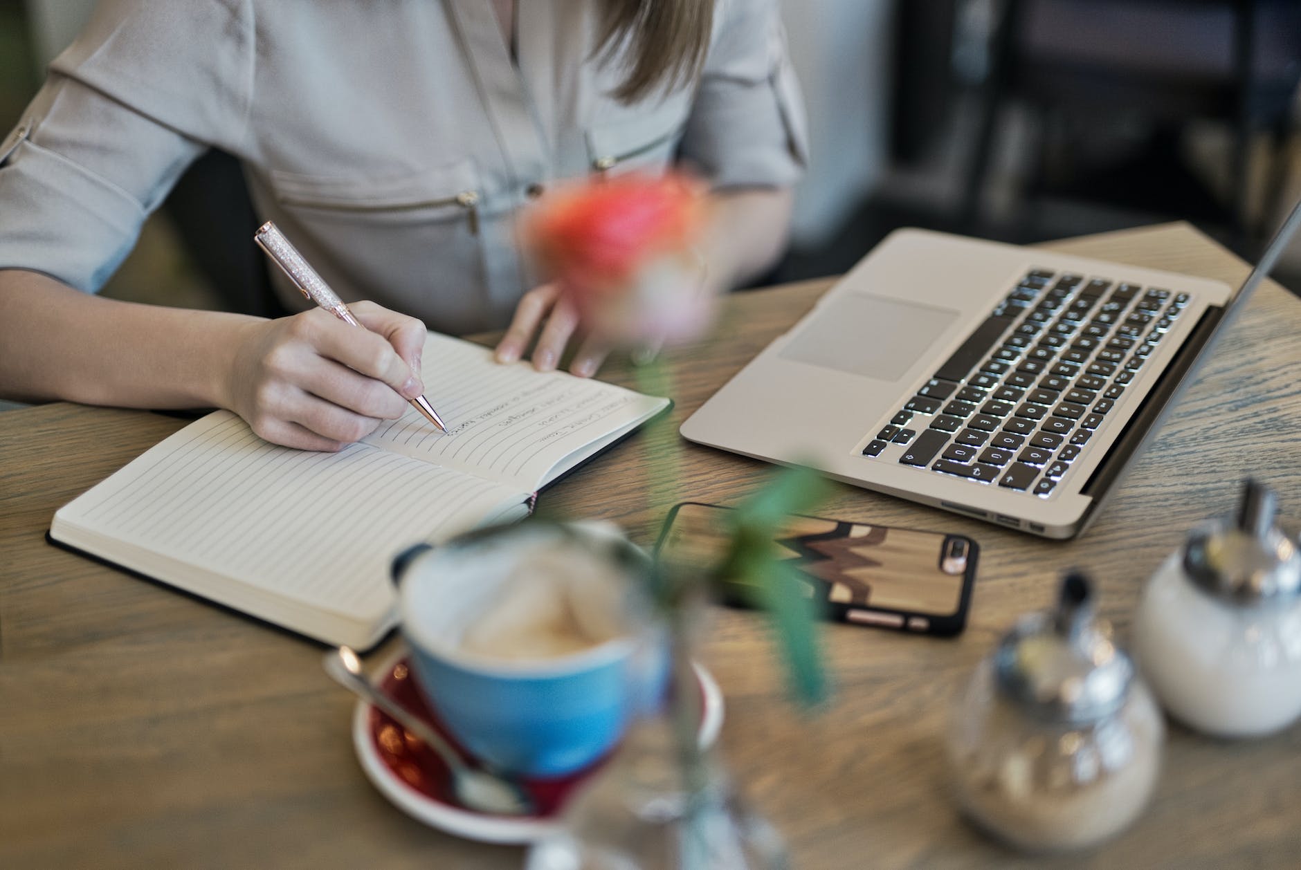 A person writing on a journal while drinking coffee and looking at a laptop next to them. 
