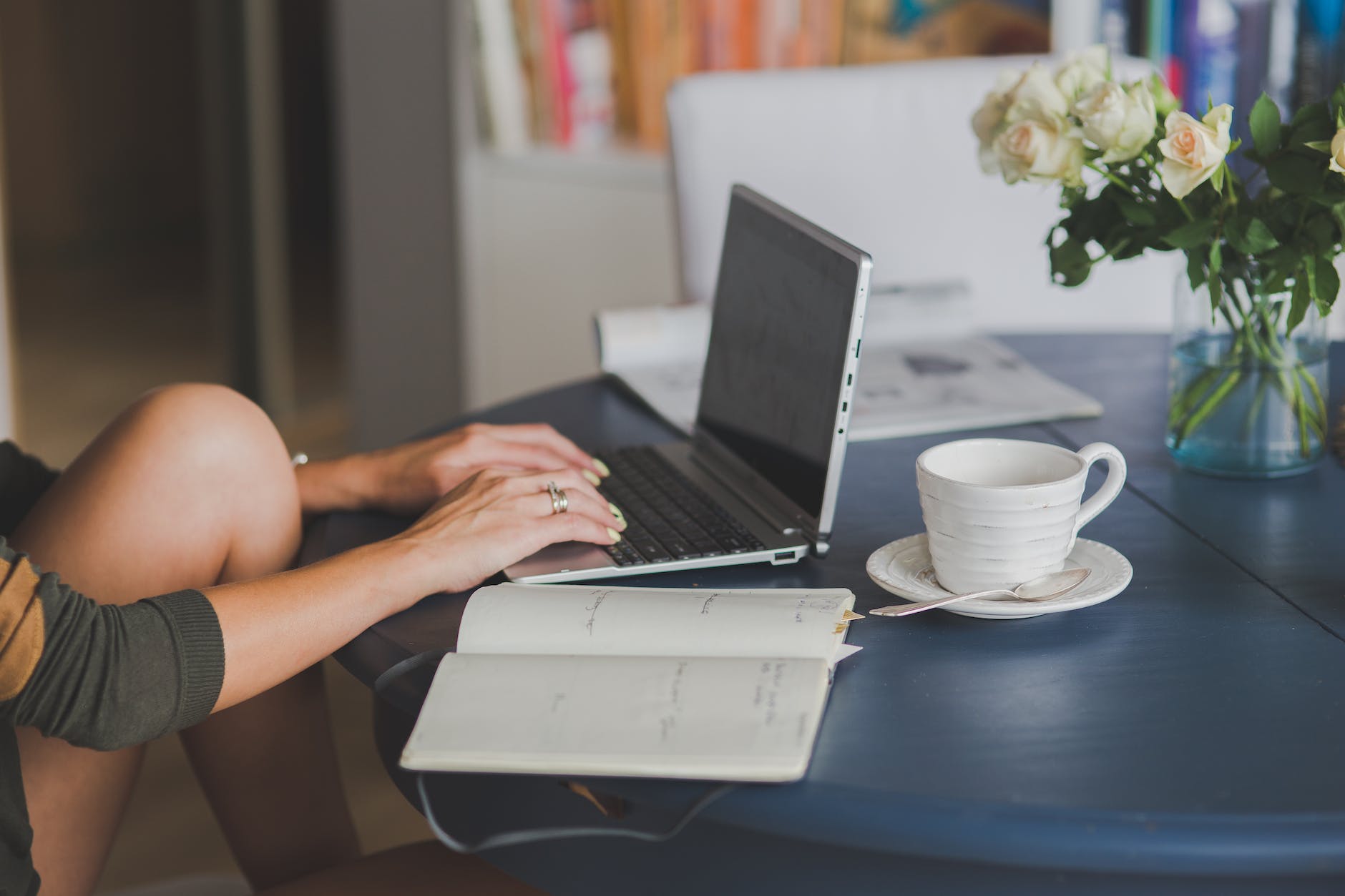 Someone using a laptop on a table kitchen, and having a cup of coffee and writing notebook next to their laptop as well as a base with white roses. 