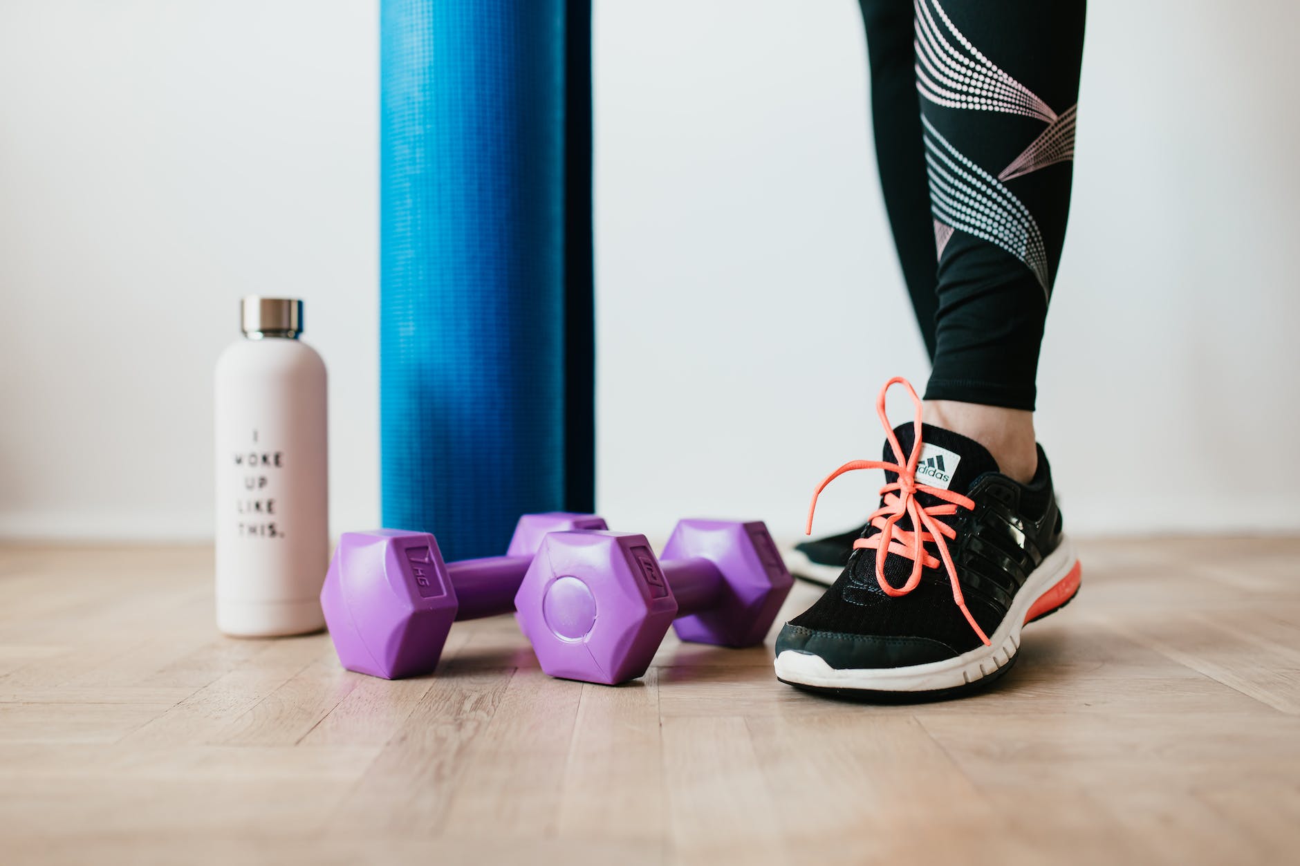 A pink water bottle next to a blue yoga mat, a pair of purple weights and workout shoes. 