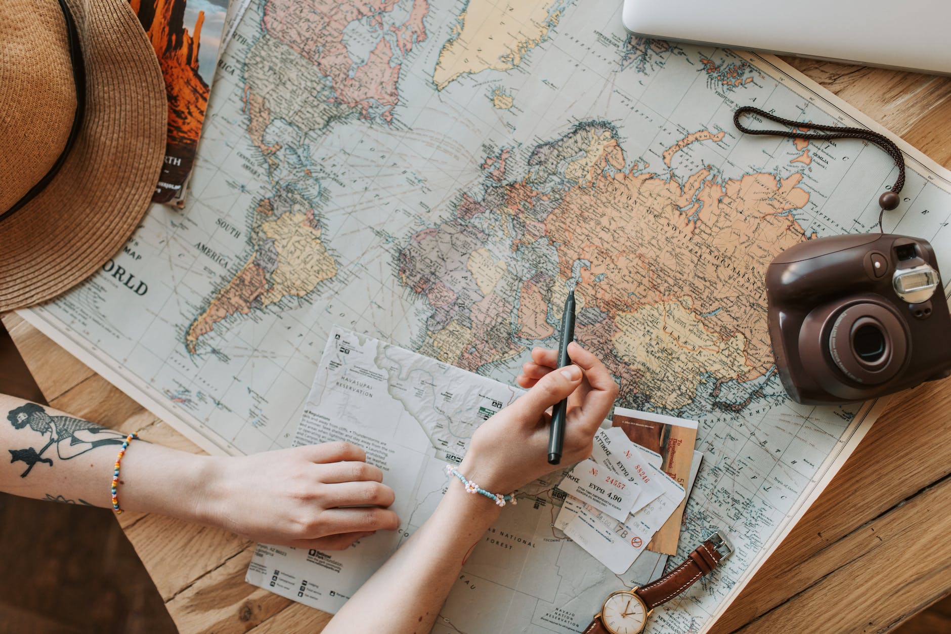 A world map laying on a table next to a polaroid camera and a watch, while a woman hands are writing on a piece of paper. 