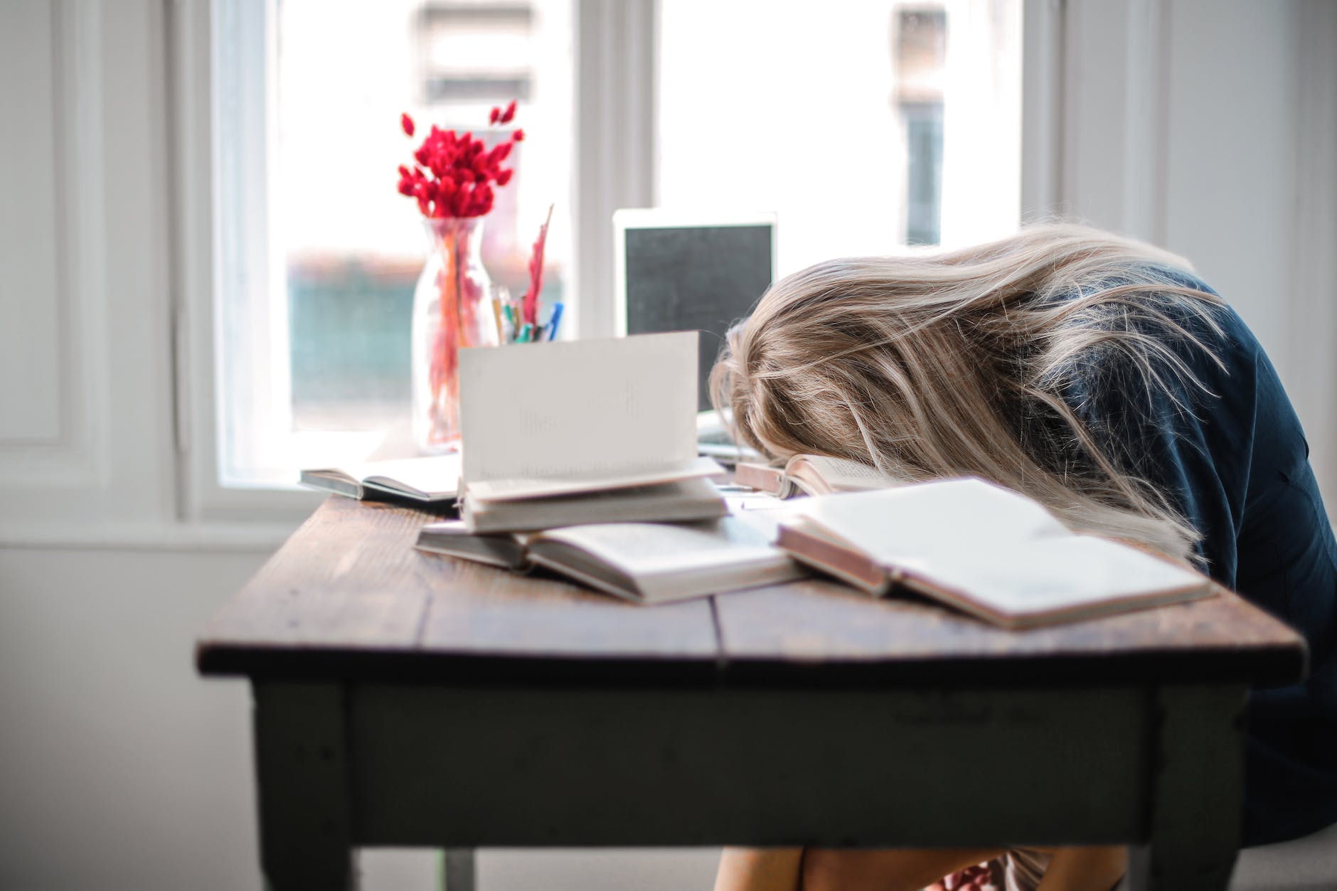 A woman feeling tired at her desk surrounded by books and her laptop. 