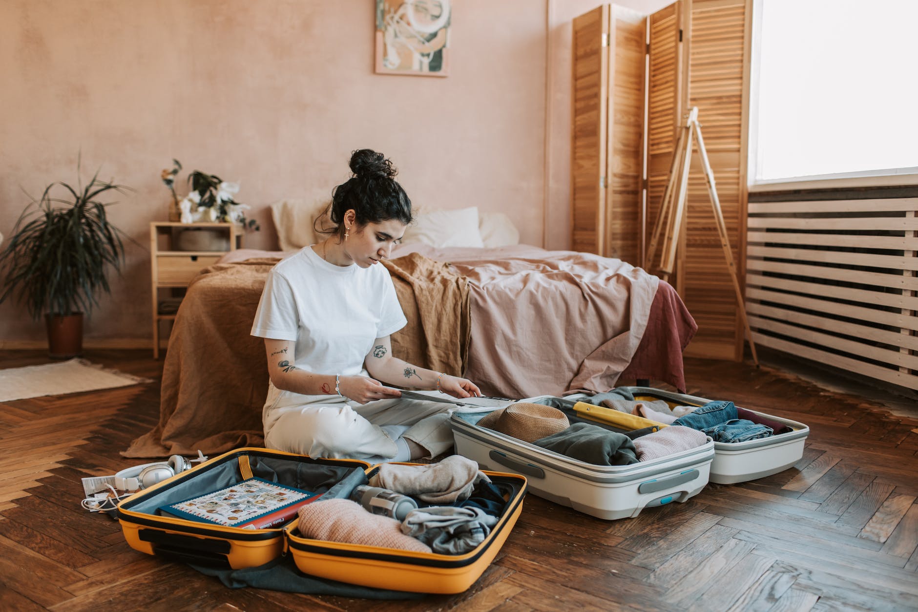 A woman wearing a white shirt and beige pants in a pink room packing clothes in two suitcases while looking into a map. 